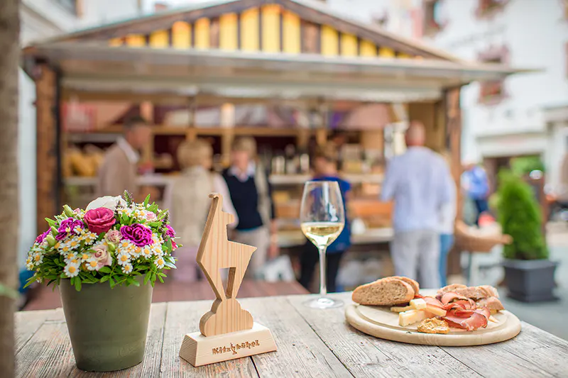 Een close-up van een tafel met een bloempot, een houten trofee, een glas witte wijn en een schaal met brood, kaas en vleeswaren. Op de achtergrond staan mensen bij een gestreepte eetkraam in de open lucht.